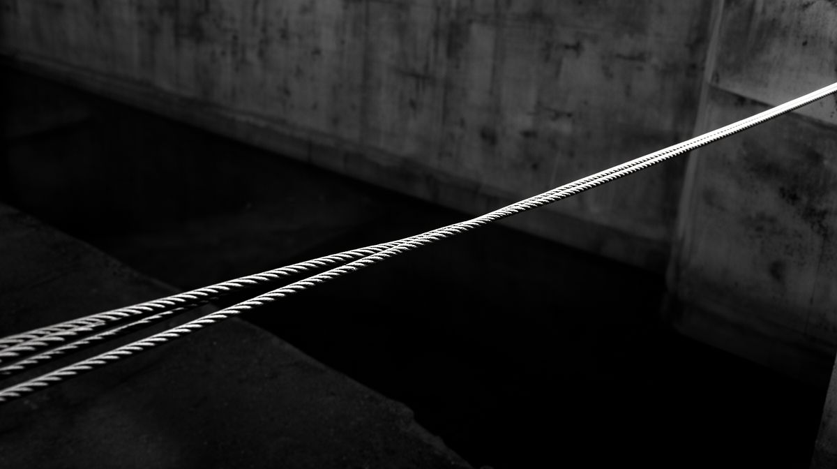 Fotografia em preto e branco de uma corda tensa esticada sobre um vazio, evocando a ideia de uma ponte frágil e imperfeita entre risco e segurança.